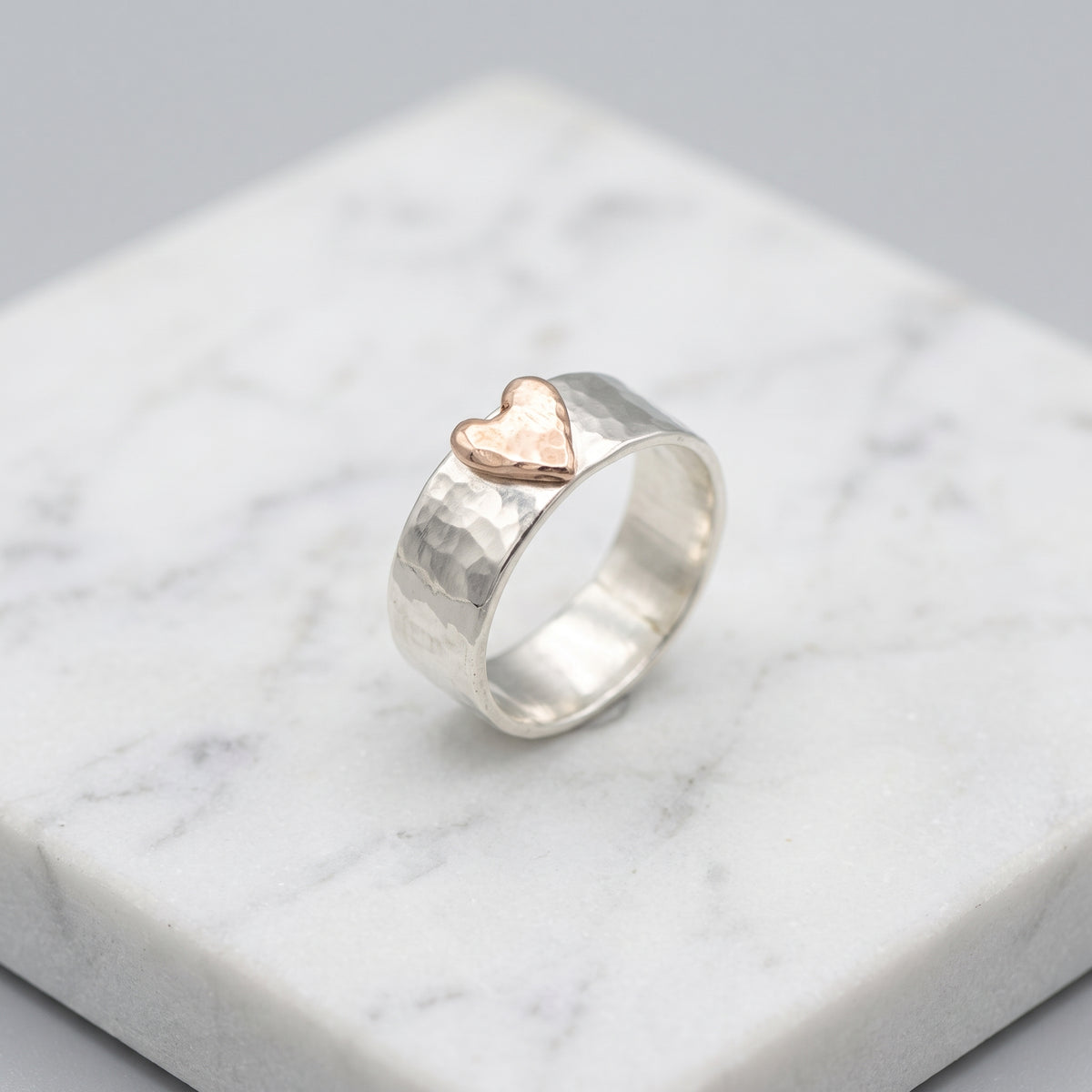 A hammered silver ring with a small rose gold heart on top, displayed on a white marble block.