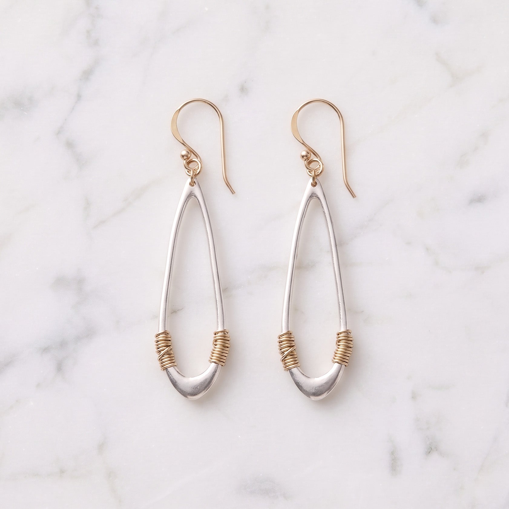 A pair of silver teardrop dangle earrings with gold wire-wrapped accents and gold fishhook ear wires, displayed on a white marble background.